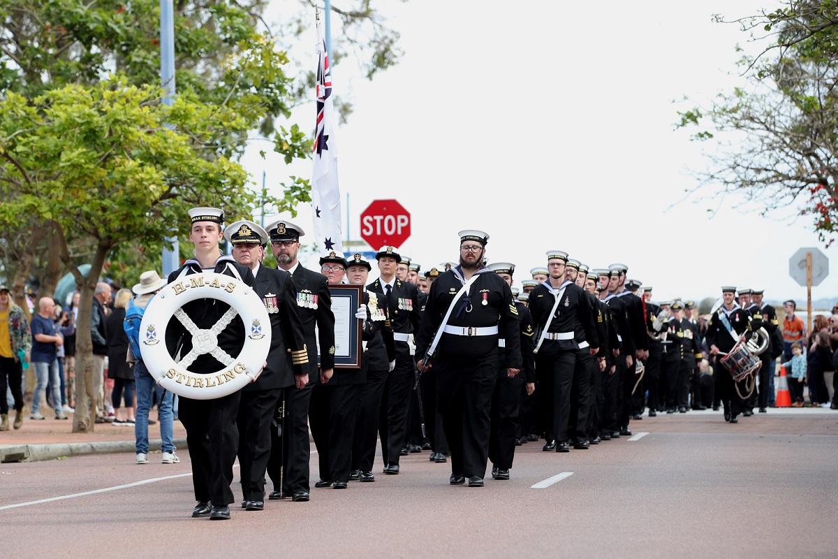 Sailors in black uniforms marching in formation during a parade, led by a member holding an HMAS Stirling life ring