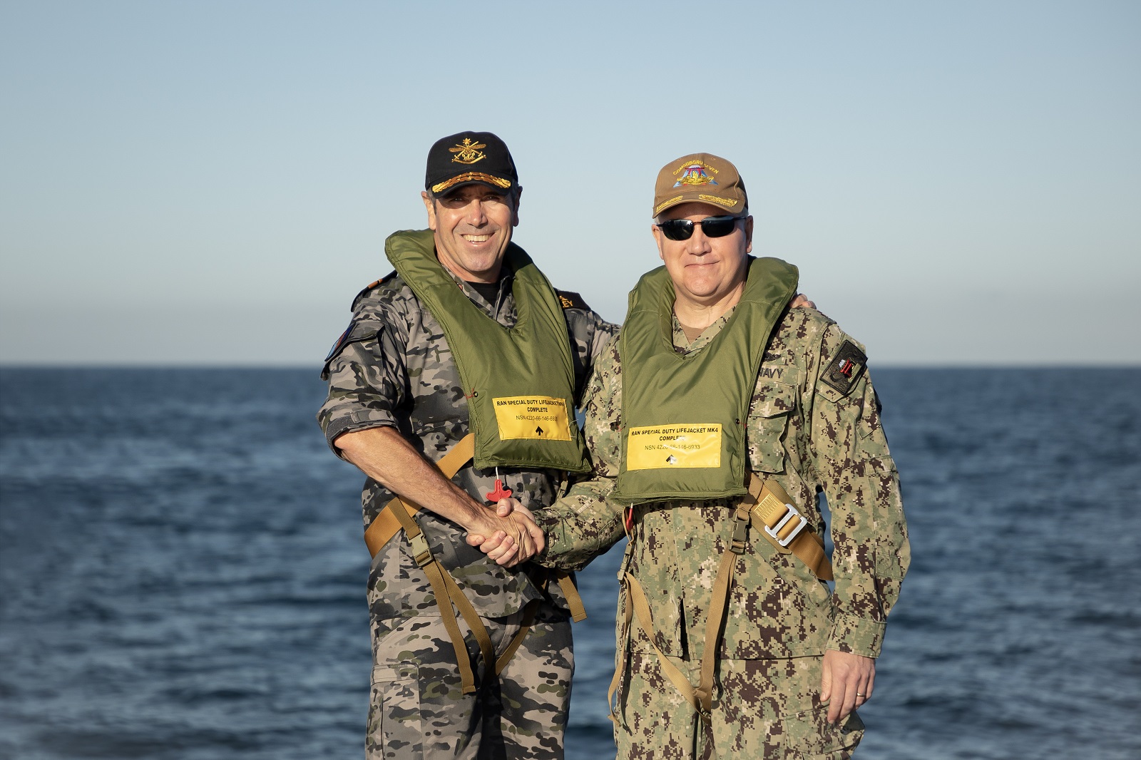 Two military personnel in camouflage uniforms and life jackets standing on a ship deck, shaking hands, with the ocean in the background