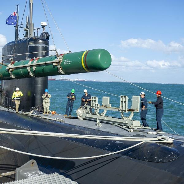 A group of people is loading a large green torpedo onto a submarine docked by the sea